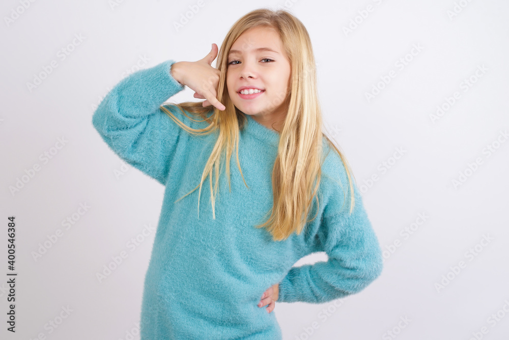 Cute Caucasian kid girl wearing blue knitted sweater against white wall imitates telephone conversation, makes phone call gesture with hands, has confident expression. Call me! Cute Caucasian kid girl wearing blue knitted sweater against white wall imitates telephone conversation, makes phone call gesture with hands, has confident expression. Call me!
