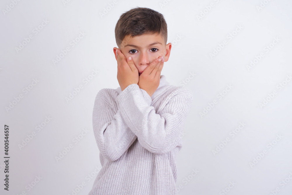 Cute Caucasian kid boy wearing knitted sweater against white wall shocked covering mouth with hands for mistake. Secret concept.
