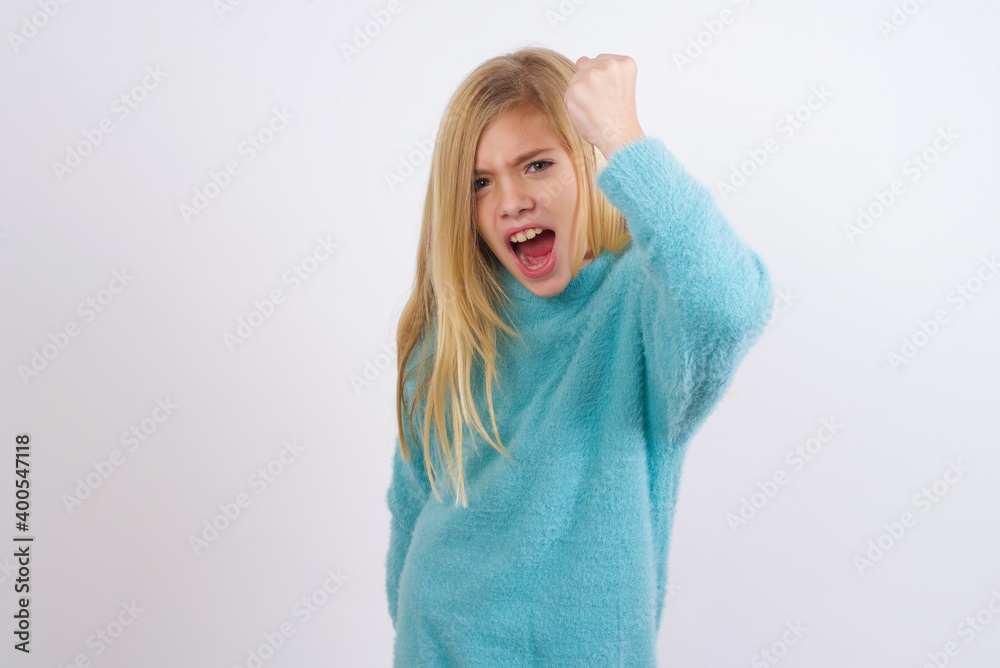 Fierce Cute Caucasian kid girl wearing blue knitted sweater against white wall holding fist in front as if is ready for fight or challenge, screaming and having aggressive expression on face. Fierce Cute Caucasian kid girl wearing blue knitted sweater against white wall holding fist in front as if is ready for fight or challenge, screaming and having aggressive expression on face.