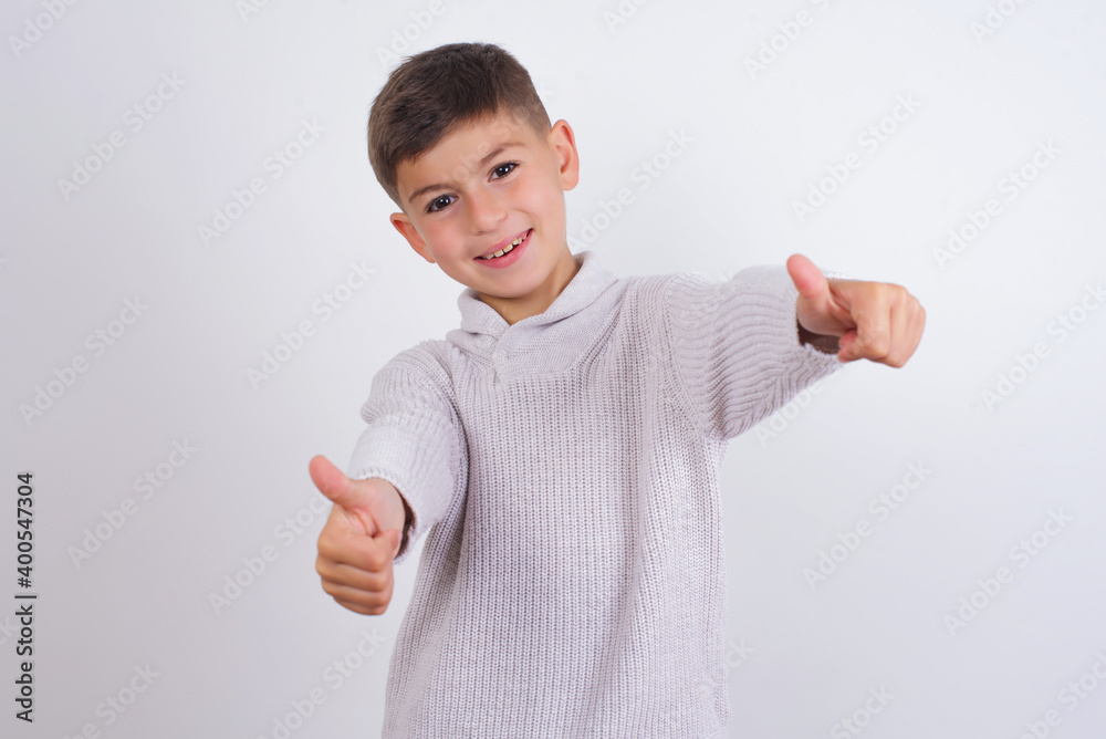 Cute Caucasian kid boy wearing knitted sweater against white wall approving doing positive gesture with hand, thumbs up smiling and happy for success. Winner gesture.