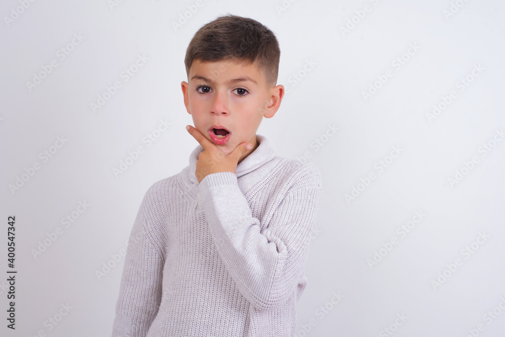 Cute Caucasian kid boy wearing knitted sweater against white wall Looking fascinated with disbelief, surprise and amazed expression with hands on chin Cute Caucasian kid boy wearing knitted sweater against white wall Looking fascinated with disbelief, surprise and amazed expression with hands on chin