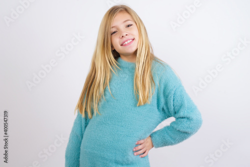 Studio shot of cheerful Cute Caucasian kid girl wearing blue knitted sweater against white wall keeps hand on hip, smiles broadly.