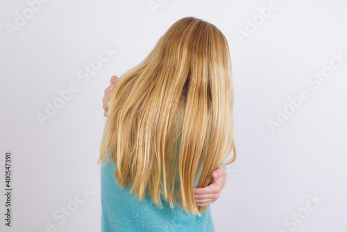 Cute Caucasian kid girl wearing blue knitted sweater against white wall hugging oneself happy and positive from backwards. Self love and self care.