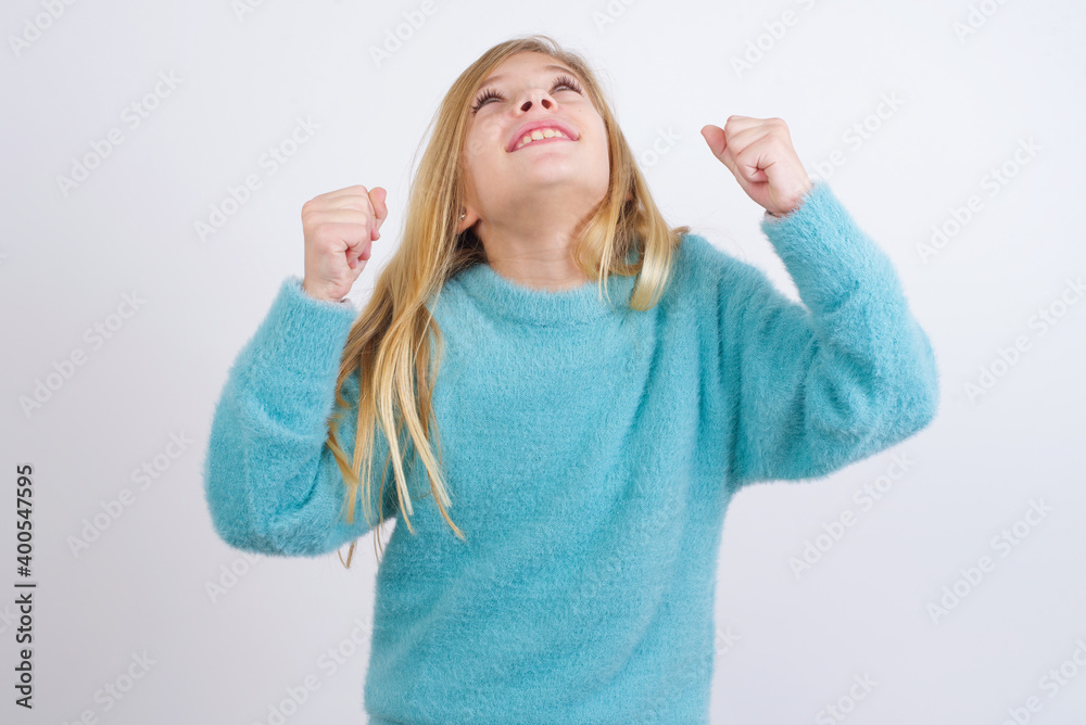 Cute Caucasian kid girl wearing blue knitted sweater against white wall having success being glad to achieve his goals. Victory and triumph concept. Cute Caucasian kid girl wearing blue knitted sweater against white wall having success being glad to achieve his goals. Victory and triumph concept.