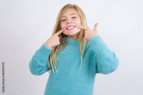 Strong healthy straight white teeth. Close up portrait of happy Cute Caucasian kid girl wearing blue knitted sweater against white wall with beaming smile pointing on perfect clear white teeth.
