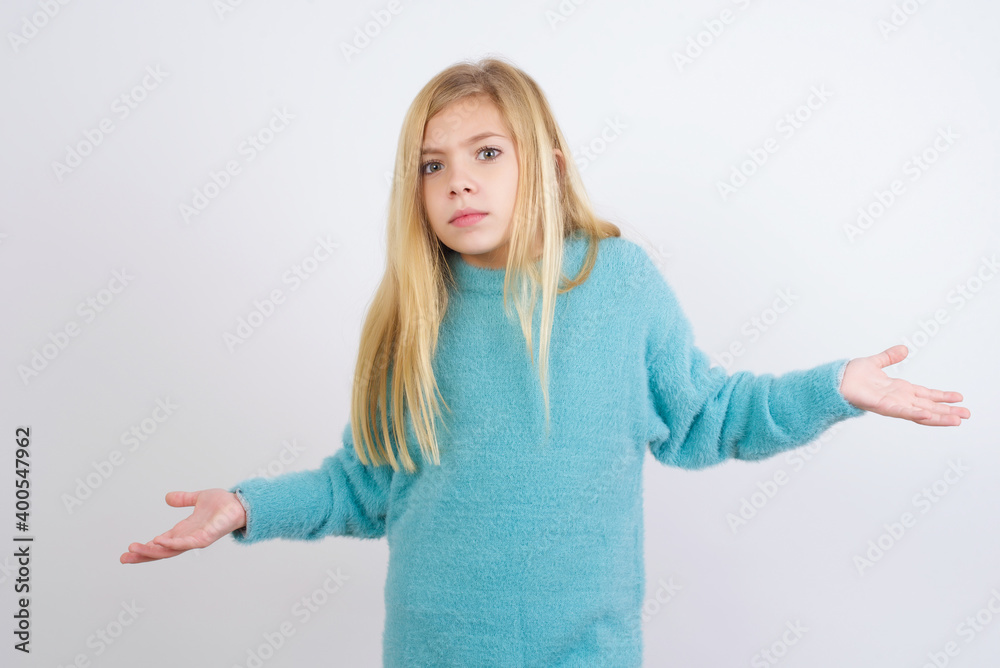 Cute Caucasian kid girl wearing blue knitted sweater against white wall looks uncertain shrugs shoulders. Cute Caucasian kid girl wearing blue knitted sweater against white wall looks uncertain shrugs shoulders.
