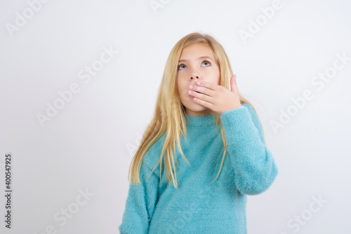 Sleepy Cute Caucasian kid girl wearing blue knitted sweater against white wall yawning with messy hair, feeling tired after sleepless night, yawning, covering mouth with palm.