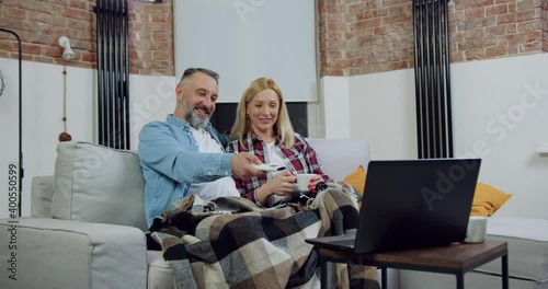 Portrait of pleasant loving carefree adult couple which relaxing on soft couch covered with blanket and enjoying interesting movie on computer photo