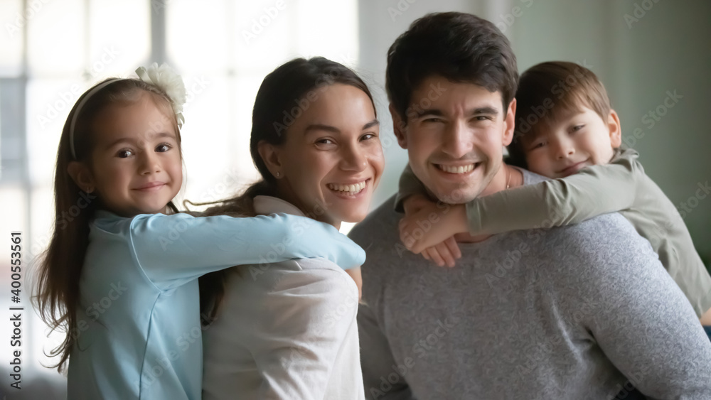 Head shot portrait smiling mother and father piggy backing adorable two kids, posing for funny family portrait at home, happy parents with little daughter and son looking at camera, having fun Head shot portrait smiling mother and father piggy backing adorable two kids, posing for funny family portrait at home, happy parents with little daughter and son looking at camera, having fun