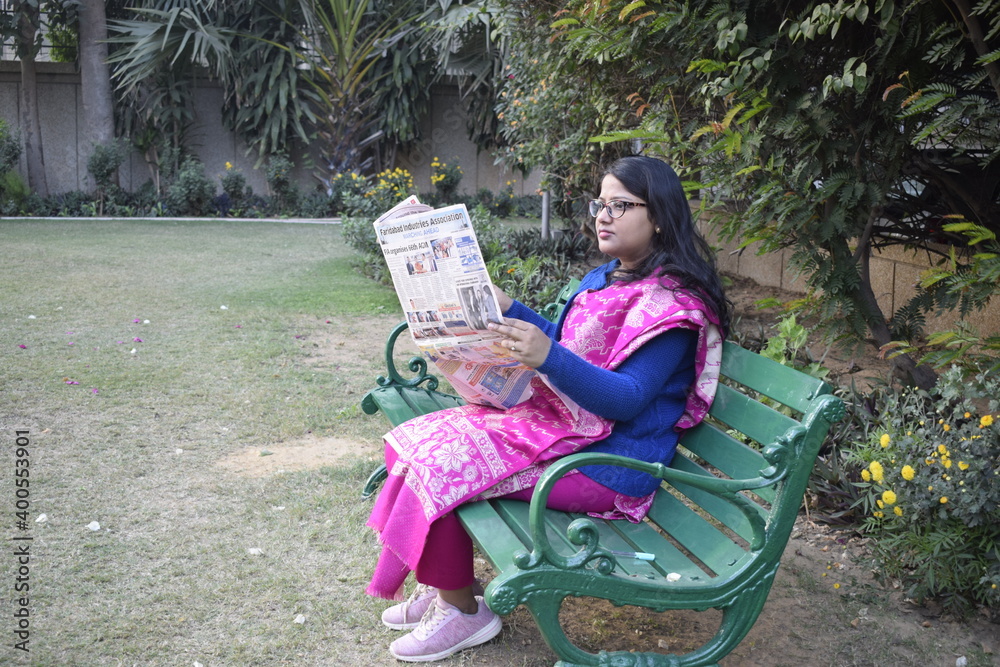Gurgaon, India 2020 : Indian girl wearing face mask sitting on a bench in a park. She is reading newspaper and enjoying sunlight during winters 