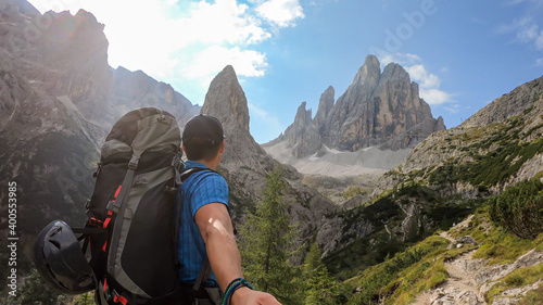 A man with big backpack taking a selfie while hiking along a narrow pathway in Italian Dolomites. There is a massive mountain with very steep and sharp slopes. Smaller mountains around. Raw landscape