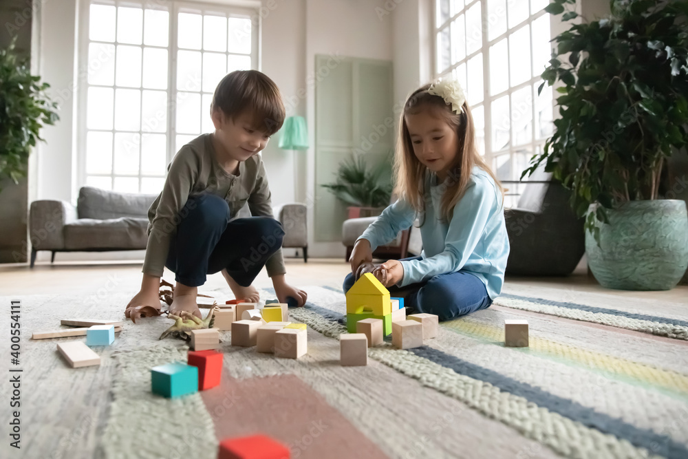 Close up little girl and boy playing with colorful wooden blocks and toy dinosaurs, sitting on warm floor with underfloor heating, curious cute sibling having fun at home together, enjoying weekend - obrazy, fototapety, plakaty 