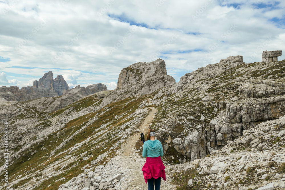 A woman hiking along a stony and narrow pathway in Italian Dolomites. There is a massive mountain in front, with very steep and sharp slopes. Some smaller mountains around. Raw and desolated landscape