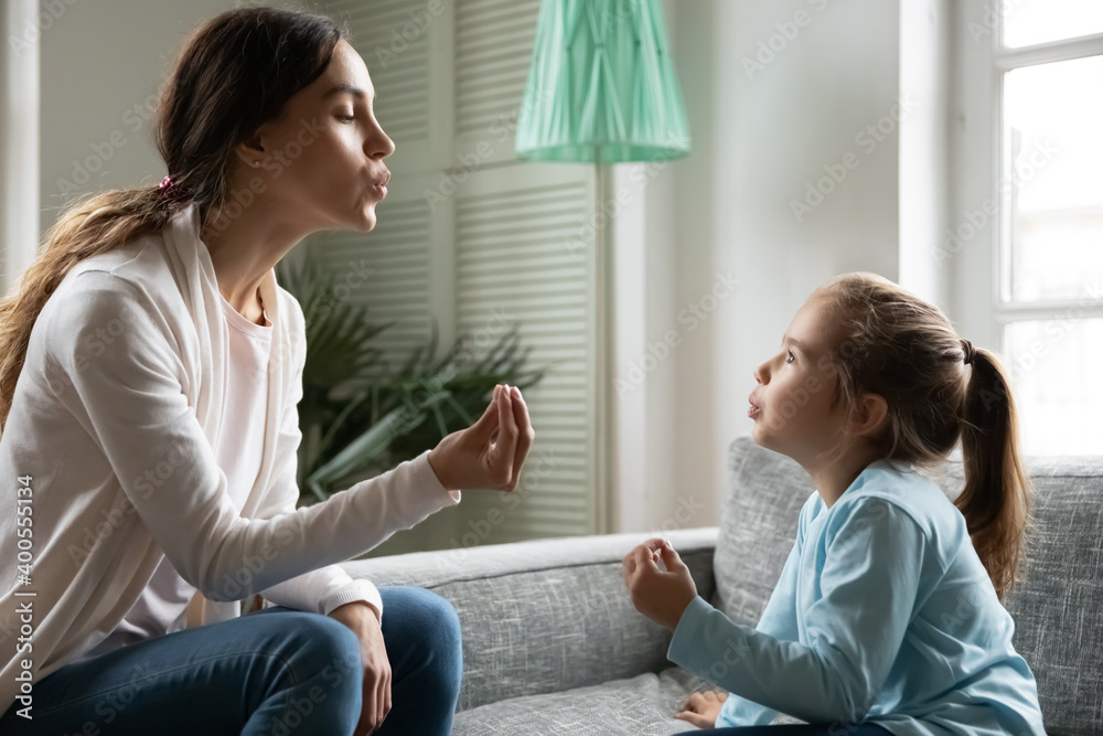 Close up little girl involved in speaking lesson with professional therapist, sitting on couch at home, caring mother teaching adorable daughter correct pronunciation, working with speech defects Close up little girl involved in speaking lesson with professional therapist, sitting on couch at home, caring mother teaching adorable daughter correct pronunciation, working with speech defects