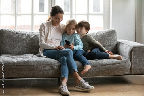 Happy mother with little son and daughter using smartphone, sitting on couch at home together, smiling mum hugging two kids, looking at phone screen, watching video, chatting or shopping online