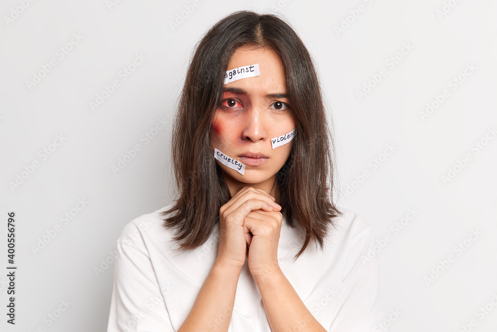 Battered bruised ethnic woman keeps hands together being afraid after abuse of violent man isolated over white background. Physically abused female with bloody eye. Stop domestic violence concept Battered bruised ethnic woman keeps hands together being afraid after abuse of violent man isolated over white background. Physically abused female with bloody eye. Stop domestic violence concept