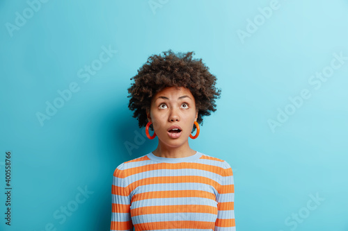 Studio shot of surprised dark skinned young woman keeps mouth opened and sees something astonishing wears casual striped jumper isolated over blue background. Human emotions and reaction concept