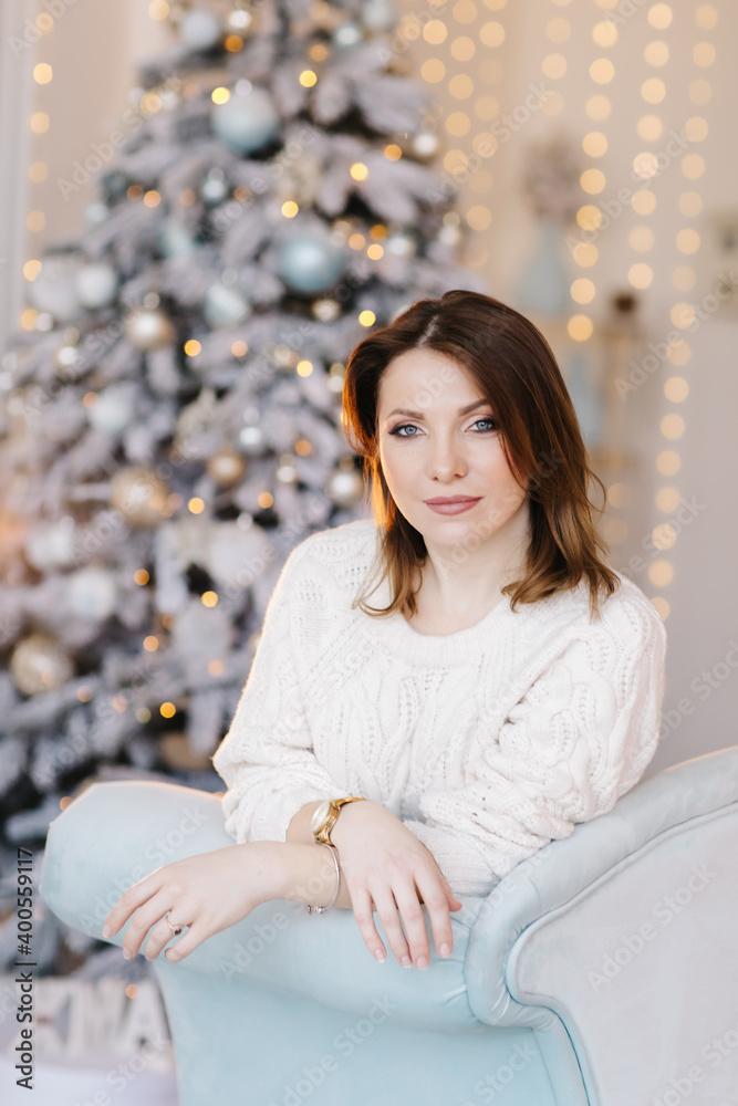 A young, attractive woman in a white sweater sits on the sofa near the Christmas tree and light
