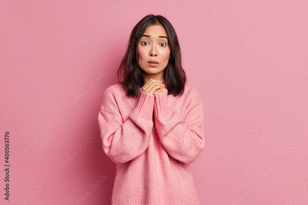 Indoor shot of young Asain brunette woman keeps hands together looks worried at camera has embarrassed face expression wears casual jumper poses against pink background. Human emotions concept Indoor shot of young Asain brunette woman keeps hands together looks worried at camera has embarrassed face expression wears casual jumper poses against pink background. Human emotions concept