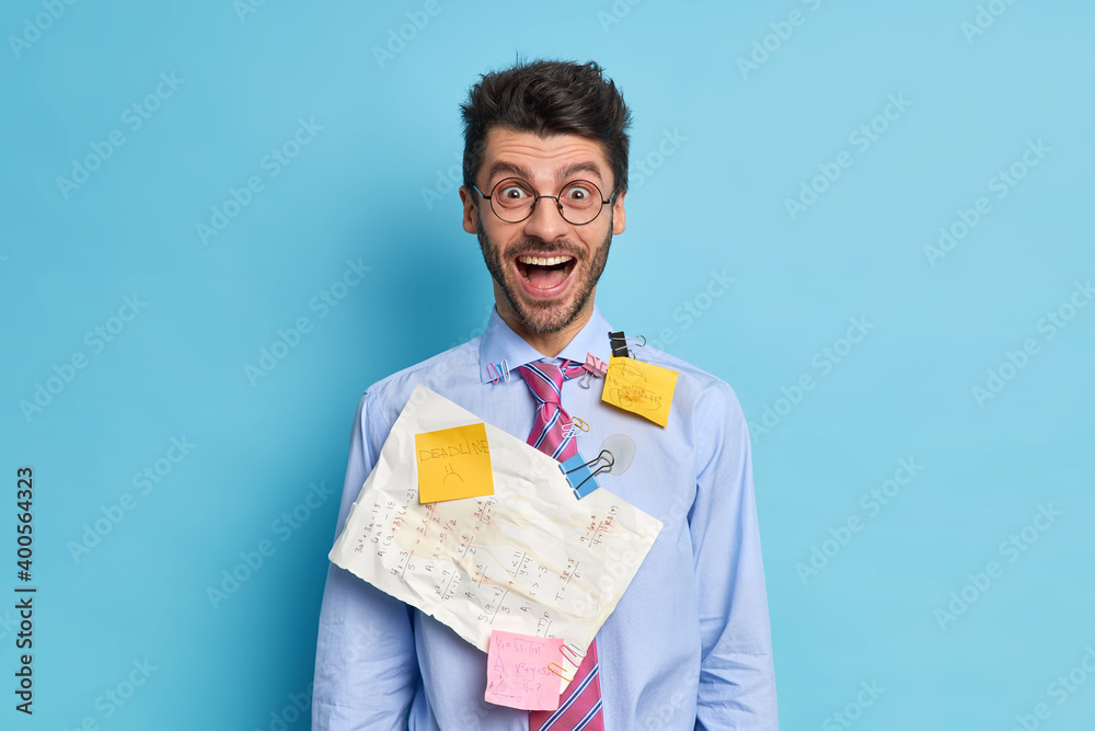 Positive male office worker has much paper work deadline for preparing research wears formal clothes and spectacles poses against blue background. Happy startuper analyzes business information Positive male office worker has much paper work deadline for preparing research wears formal clothes and spectacles poses against blue background. Happy startuper analyzes business information