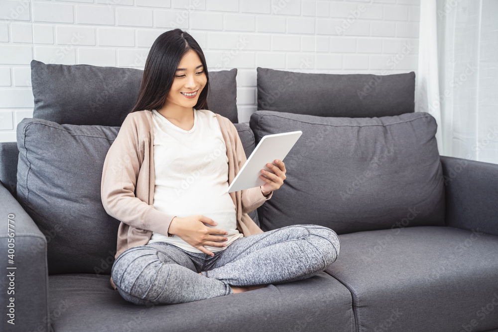 beautiful asian pregnant woman smiling using smart tablet device watching video placing hands on baby lump, sitting on sofa relaxing resting in living room with brick texture wall and white curtains beautiful asian pregnant woman smiling using smart tablet device watching video placing hands on baby lump, sitting on sofa relaxing resting in living room with brick texture wall and white curtains