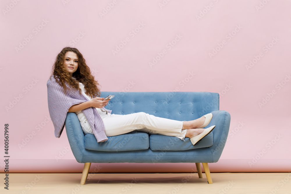 Beautiful curly-haired girl in casual 
white clothes and purple sweater sits on blue sofa. Pastel trendy pink background. Portrait of smiling young woman with smartphone in hands.