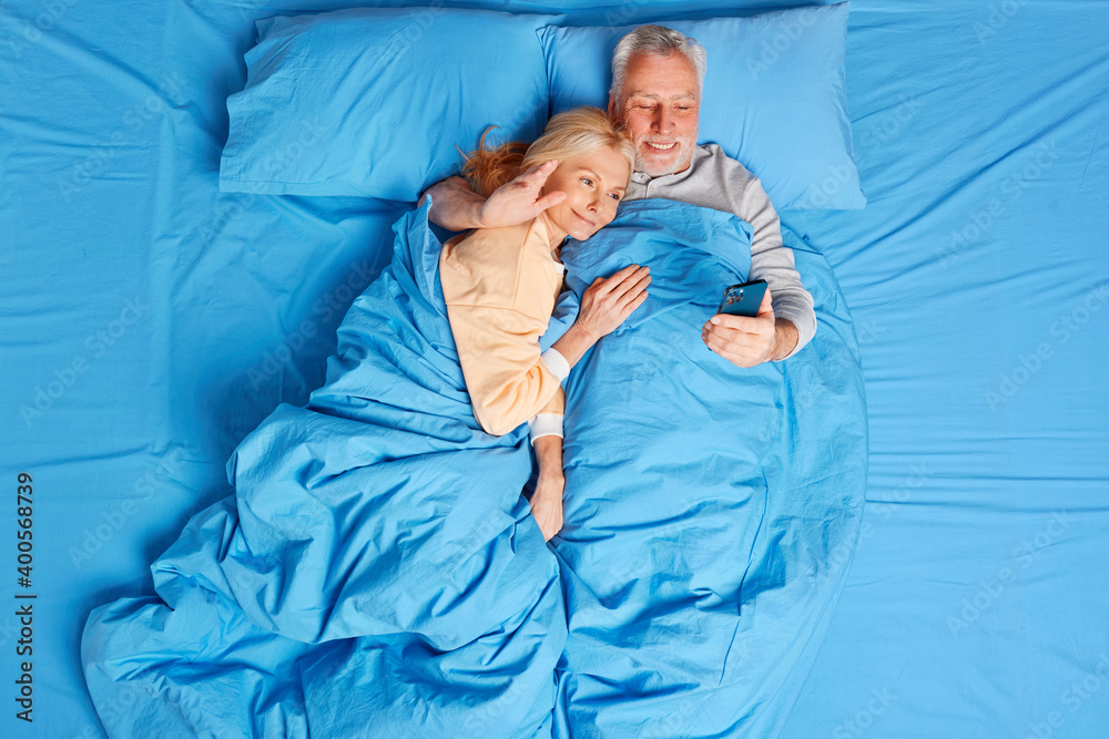 Top view of glad mature woman and man lay in bed talk distantly with