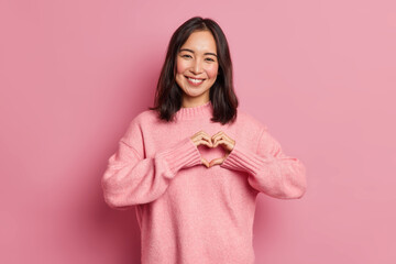 Attractive brunette young Asian woman feels happy and romantic shapes heart gesture expresses tender feelings wears casual jumper poses against pink background. People affection and care concept