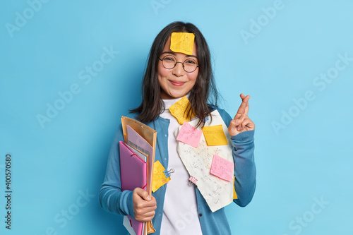 Exam preparation concept. Pleased Asian woman crosses fingers believes in good luck makes financial research prepares for brainstroming meeting poses satisfied against blue studio background