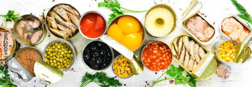 Food background in tin cans. Canned vegetables, beans, fish and fruits on a white wooden background. Top view.