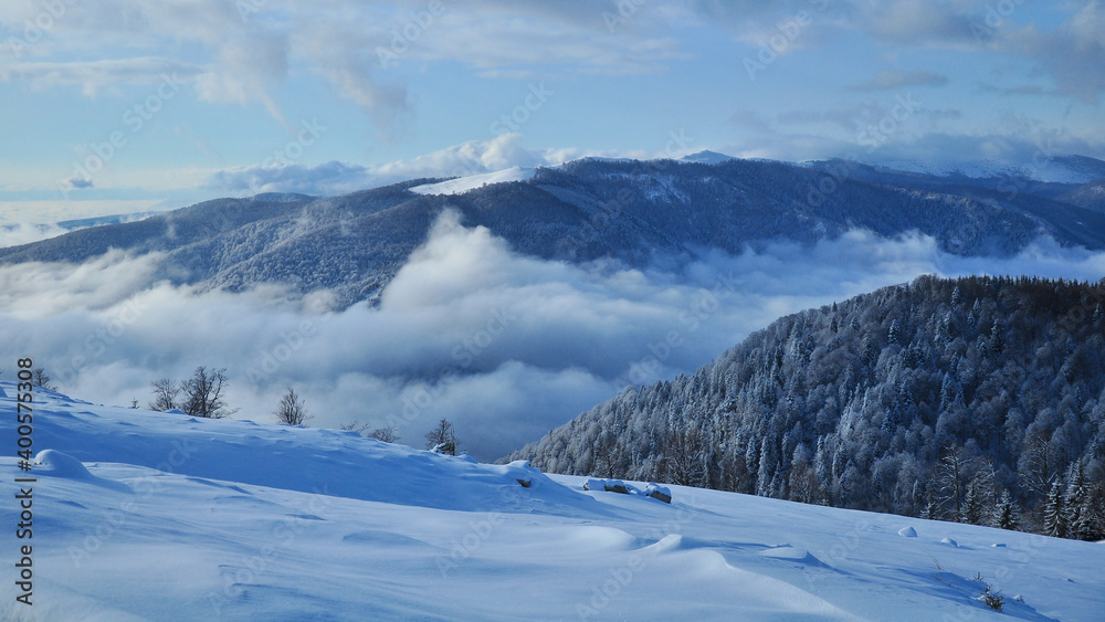 Snowed mountain crest standing above the thick layer of clouds and fog covering the forested valley below. Panorama made in Parang Mountains, Carpathia, Romania.