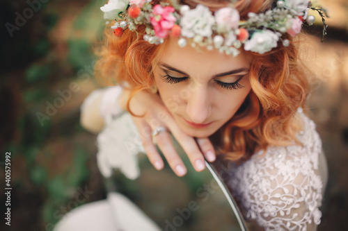 beautiful young red-haired bride in the forest with a floral wreath on her head and mirror . woman in long white dress outdoors on summer day. wedding day