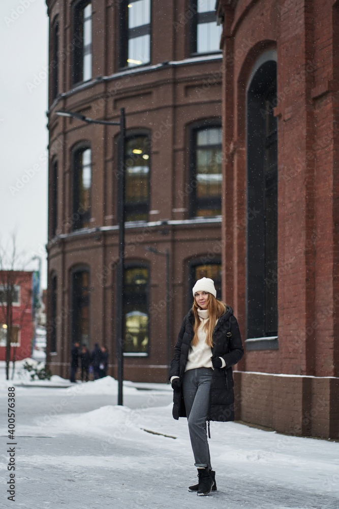 beautiful young woman with long hair, in a dark jacket, white hat and a sweater with a high neck against the background of a loft building made of red brick with tall black windows. snowing beautiful young woman with long hair, in a dark jacket, white hat and a sweater with a high neck against the background of a loft building made of red brick with tall black windows. snowing