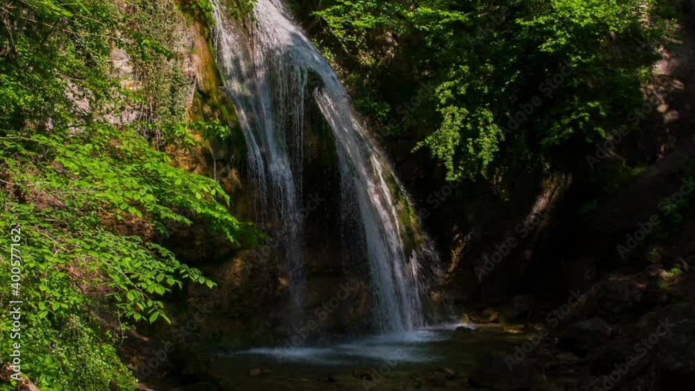 Slow-motion shooting of a waterfall in the forest on a sunny day. The sun's rays illuminate the cascade of water falling with foam from the cliff. A forest landscape that gives you peace and coolness Slow-motion shooting of a waterfall in the forest on a sunny day. The sun's rays illuminate the cascade of water falling with foam from the cliff. A forest landscape that gives you peace and coolness
