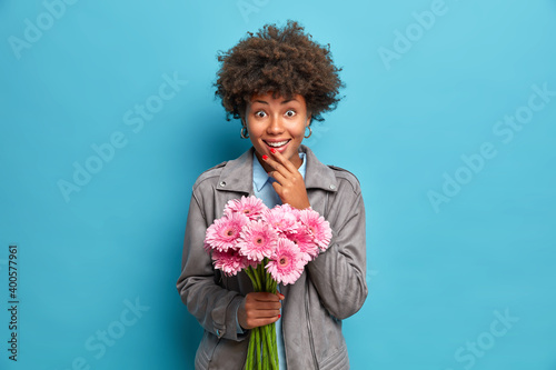 Fashionable young Afro American woman receives bouquet of pink gerbera flowers from loving boyfiend during date wears stylish grey jacket poses against blue background. Happy Womens Day concept