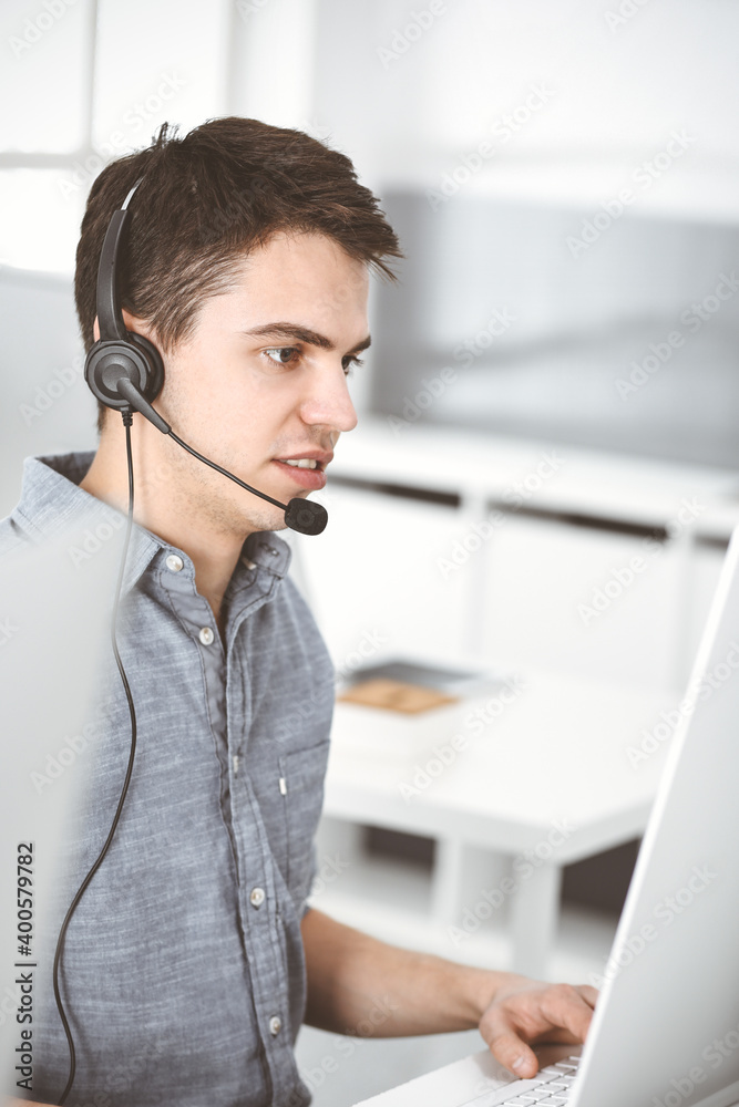 Casual dressed young man using headset and computer while talking with customers online. Call center, business concept Casual dressed young man using headset and computer while talking with customers online. Call center, business concept