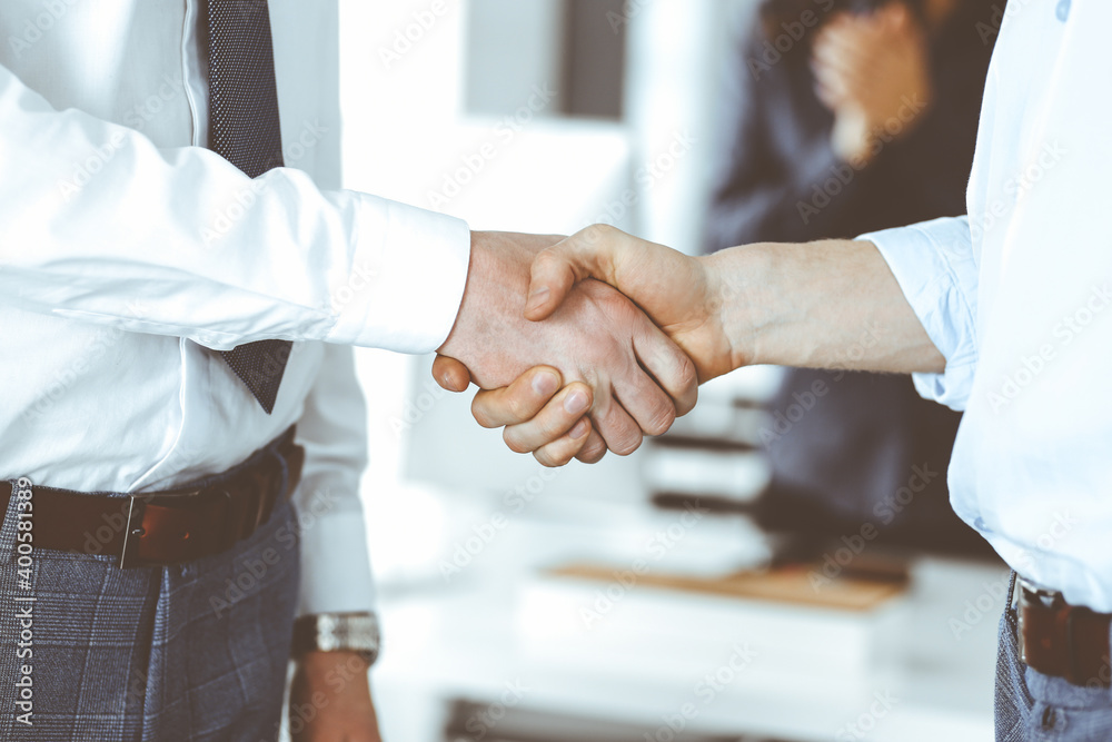Fototapeta premium Two businessmen are shaking hands in office, close-up. Happy and excited business woman stands with raising hands at the background. Business people concept