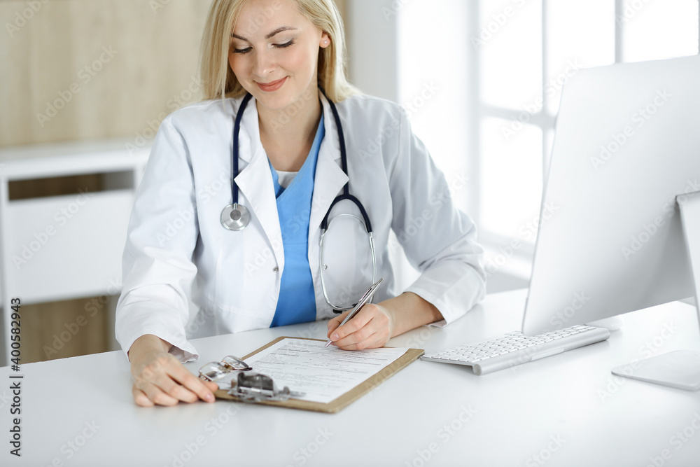 Woman-doctor at work while sitting at the desk in hospital or clinic. Blonde cheerful physician filling up medication history record form Woman-doctor at work while sitting at the desk in hospital or clinic. Blonde cheerful physician filling up medication history record form