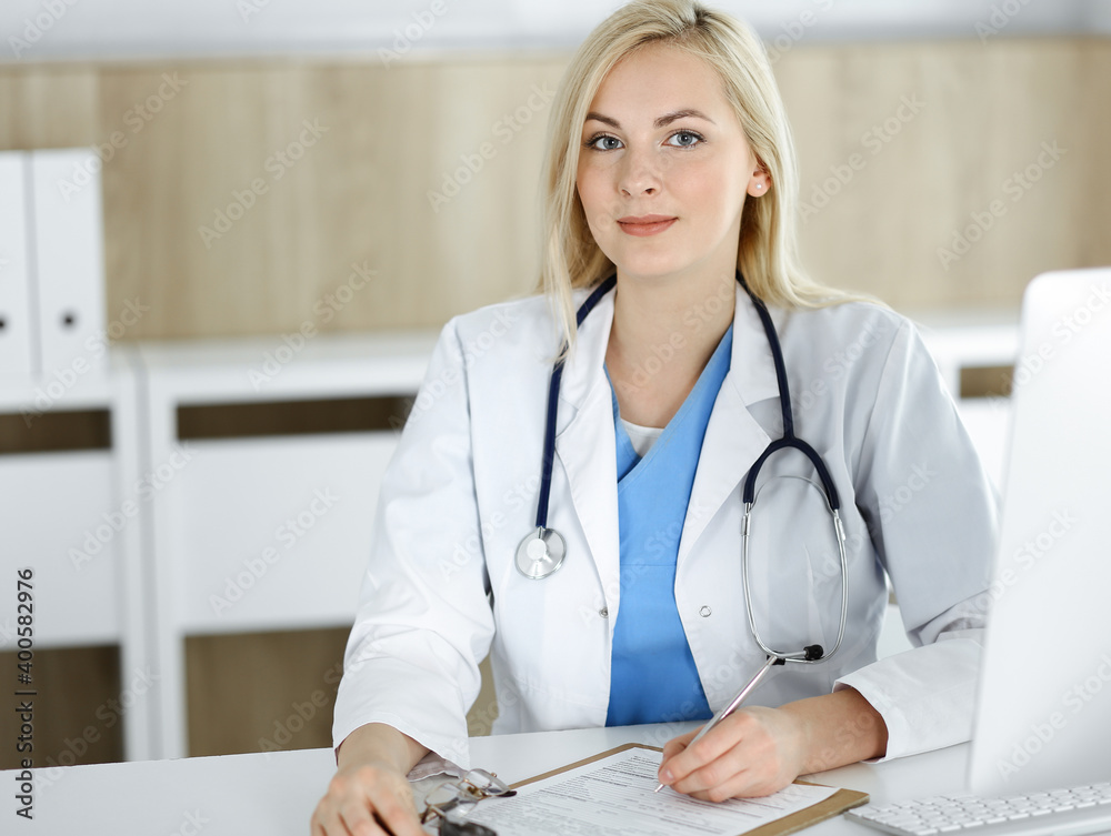 Portrate of woman-doctor at work while sitting at the desk in clinic. Blonde cheerful physician filling up medical form or prescription Portrate of woman-doctor at work while sitting at the desk in clinic. Blonde cheerful physician filling up medical form or prescription