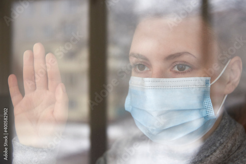 A woman in a medical mask looks out the window, self-isolation during the coronavirus.