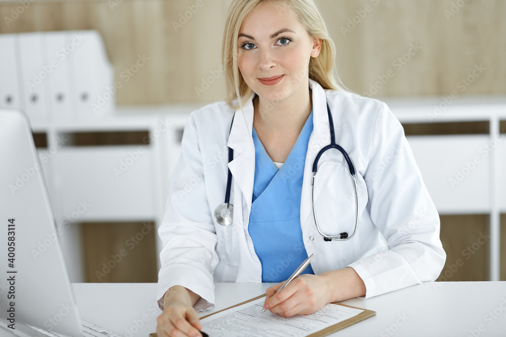 Portrate of woman-doctor at work while sitting at the desk in clinic. Blonde cheerful physician filling up medical form or prescription Portrate of woman-doctor at work while sitting at the desk in clinic. Blonde cheerful physician filling up medical form or prescription