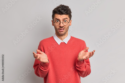 Indignant puzzled man raises palms and faces difficult choice feels hesitant wears round transparent glasses red jumper stands displeased against grey background. Human perception attitude concept photo