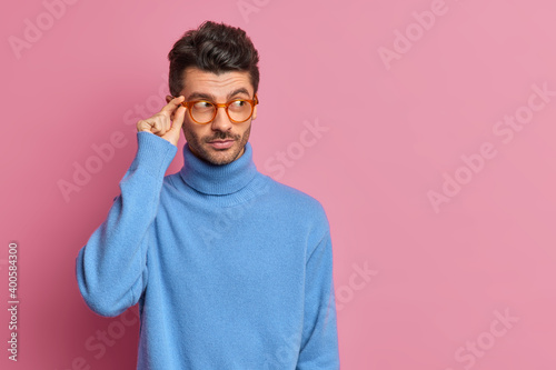 Studio shot of thoughtful European man concentrated aside pensively wears transparent glasses and blue turtleneck thinks about question has pensive look poses against pink background blank copy space