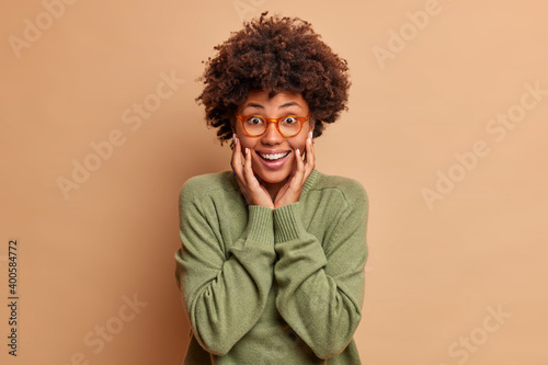 Joyful woman with Afro hair keeps hands on cheeks smiles happily looks gladfully at camera wears optical glasses and sweater isolated over brown background. Positive emotions and human reactions