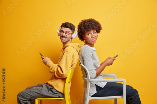 Dissatisfied millennial woman and man sit back to each other on chairs clench teeth use mobile phones check social networks news online use new app annoyed by spam isolated over yellow wall. © wayhome.studio 