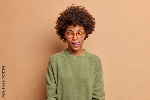 Photo of Afro American teenage girl makes funny grimace crosses eyes and sticks out tongue wears basic sweater poses against brown background. Playful woman entertains child. Face expressions concept © wayhome.studio 