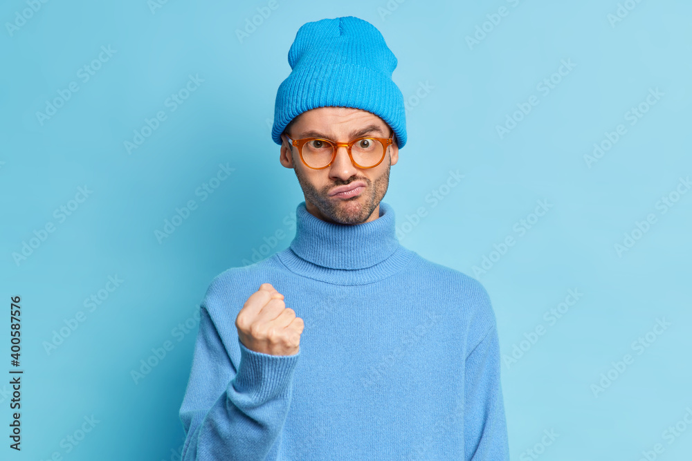 Indoor shot of displeased unshaven man clenches fist expresses anger and asks not to bother him wears blue hat spectacles turtleneck isolated over blue background looks with hateful expression