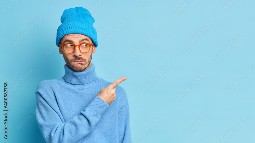 Puzzled man teenager purses lips wears spectacles blue casual poloneck and hat points away on copy space surprised by unexpected offer poses against blue background. Place your promotion here Puzzled man teenager purses lips wears spectacles blue casual poloneck and hat points away on copy space surprised by unexpected offer poses against blue background. Place your promotion here