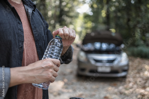 Man open water bottle in front of the open hood of a broken car on the road in the forest hot weather day. Car breakdown concept. photo