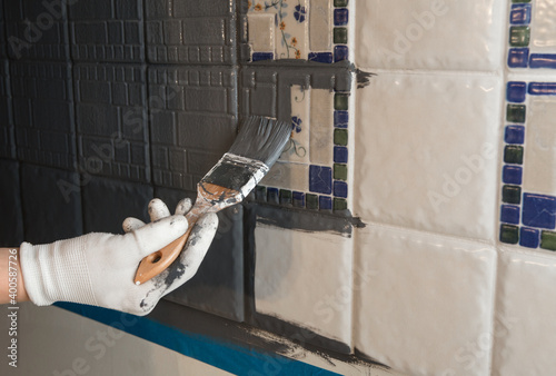 Repainting old dated kitchen ceramic tile back wall with modern gray chalk paint indoors at home. Giving old kitchen new look concept. Hand holding a paint brush tool. © FotoHelin